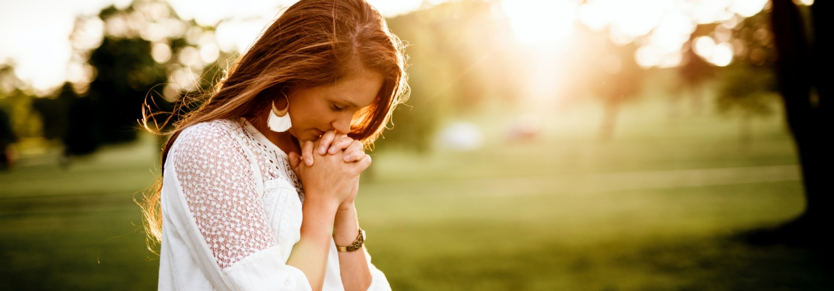 Woman with clasped hands standing outside under a tree praying and being thoughtful