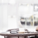 A man sits at a table in a cafe, working on his laptop while smiling. He has a cup of coffee and headphones around his neck.