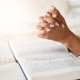 Prayer, bible and hands praying for religion, hope or help, spiritual or faith in home. God, christian and female or woman worship Jesus Christ or Holy Spirit with catholic text or book for peace